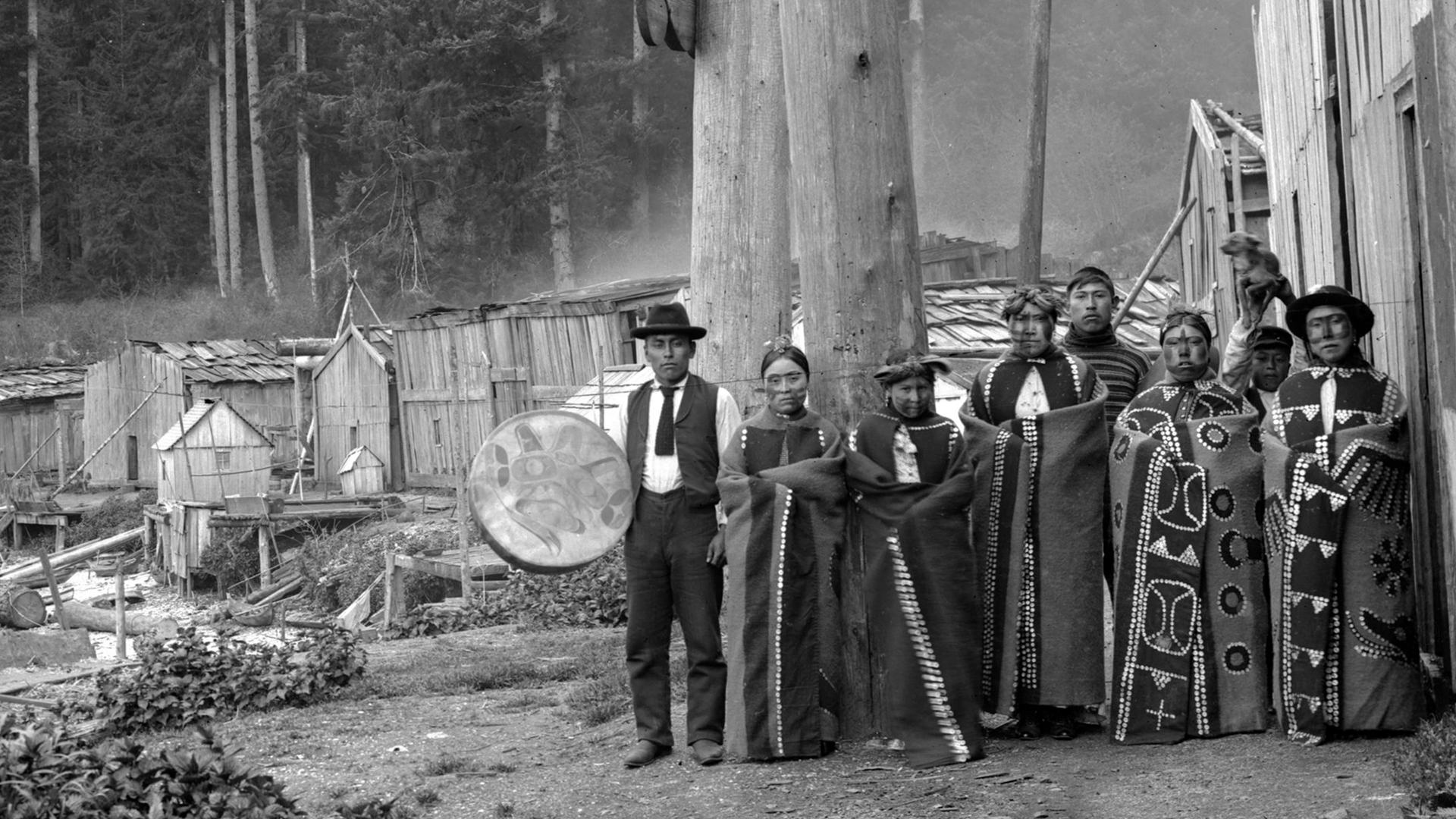 Eight members of the Kwakiutl First Nations are photographed outdoors in traditional dress and face paint, circa. 1895 – 1898. The man on the leftmost side holds a large drum.