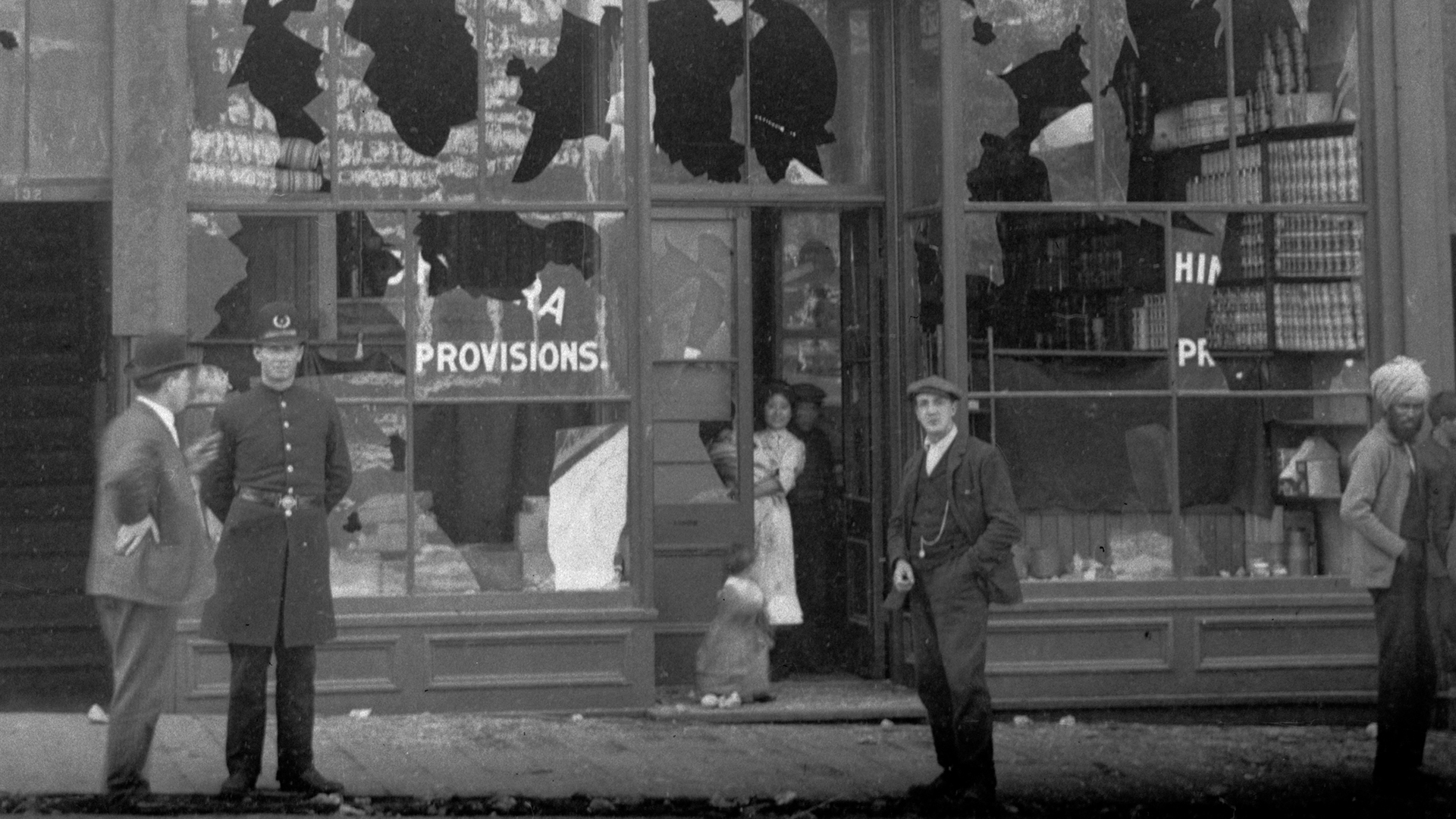Following the Vancouver anti-Asian riot in September 1907, the storefront of Nishimura Masuya’s grocery store is damaged with shattered windows. Onlookers include a police officer, an Asian woman in the doorway of the store, and a South Asian man.