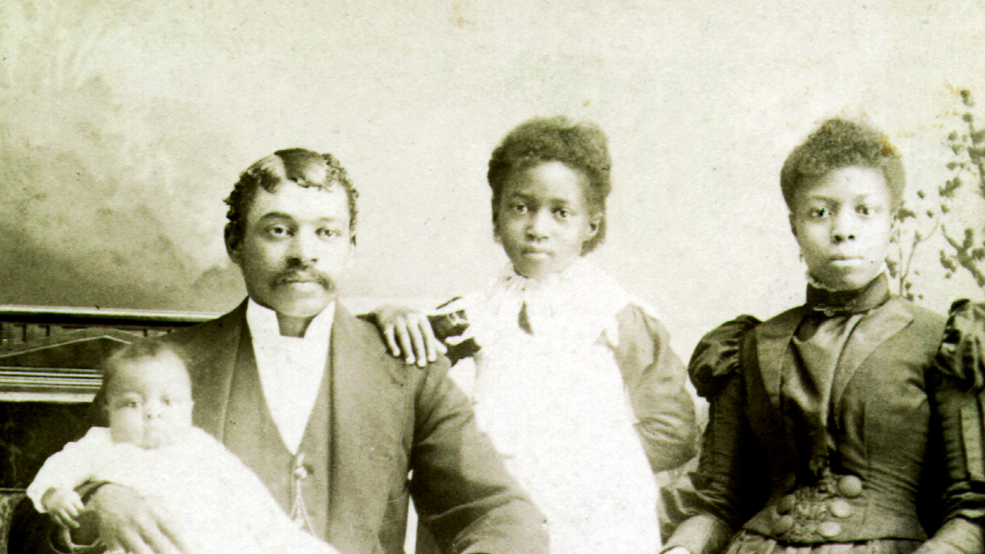 A Black couple and their two young children – one school-aged daughter and an infant – pose for a family portrait, circa. 1850s. They are dressed in typical Victorian-era fashion. 
