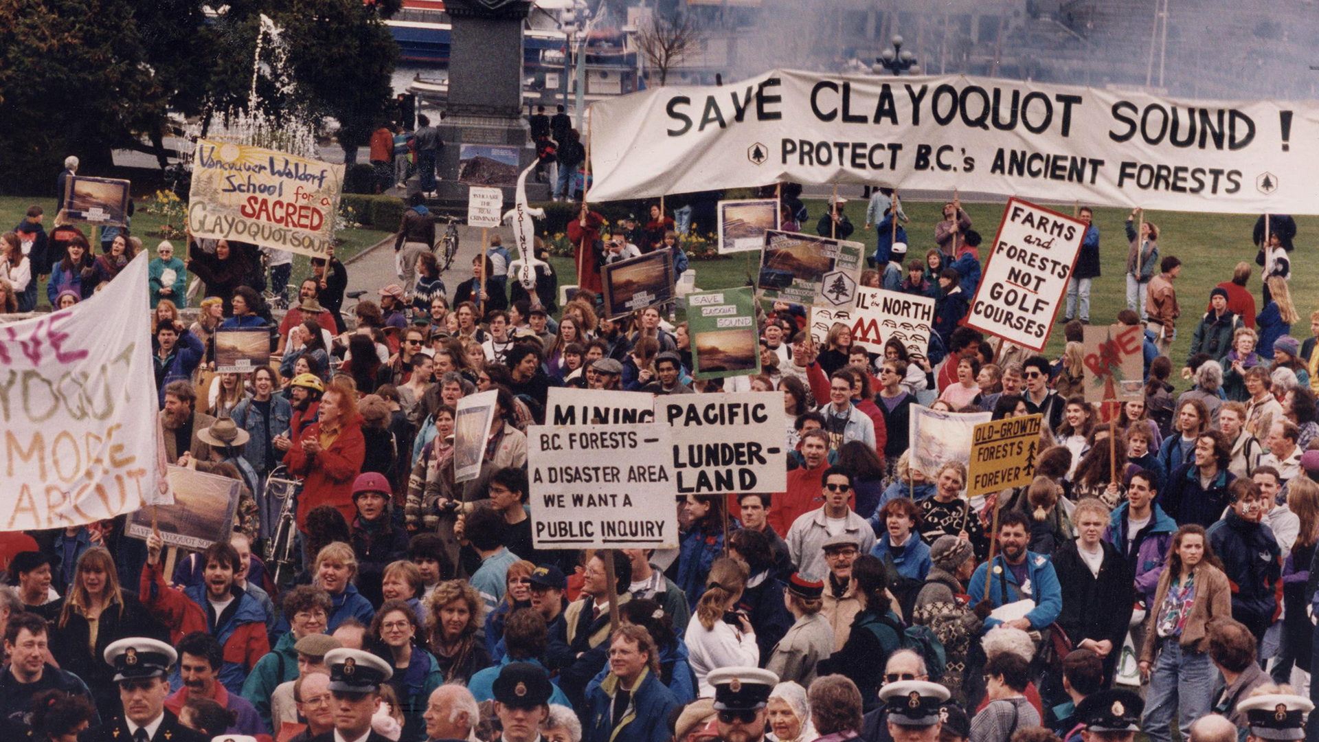 A protest to protect the forests of Clayoquot Sound, BC takes place in Victoria in March 1993. A large group of people holding signs and banners gather outdoors. 